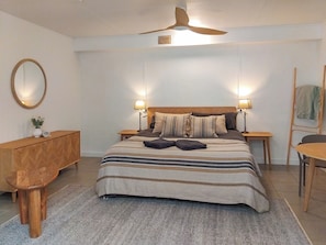 Ein Schlafzimmer mit einem Bett, zwei Nachttischen, einer Holzbank, einem runden Spiegel und einem Deckenventilator.
