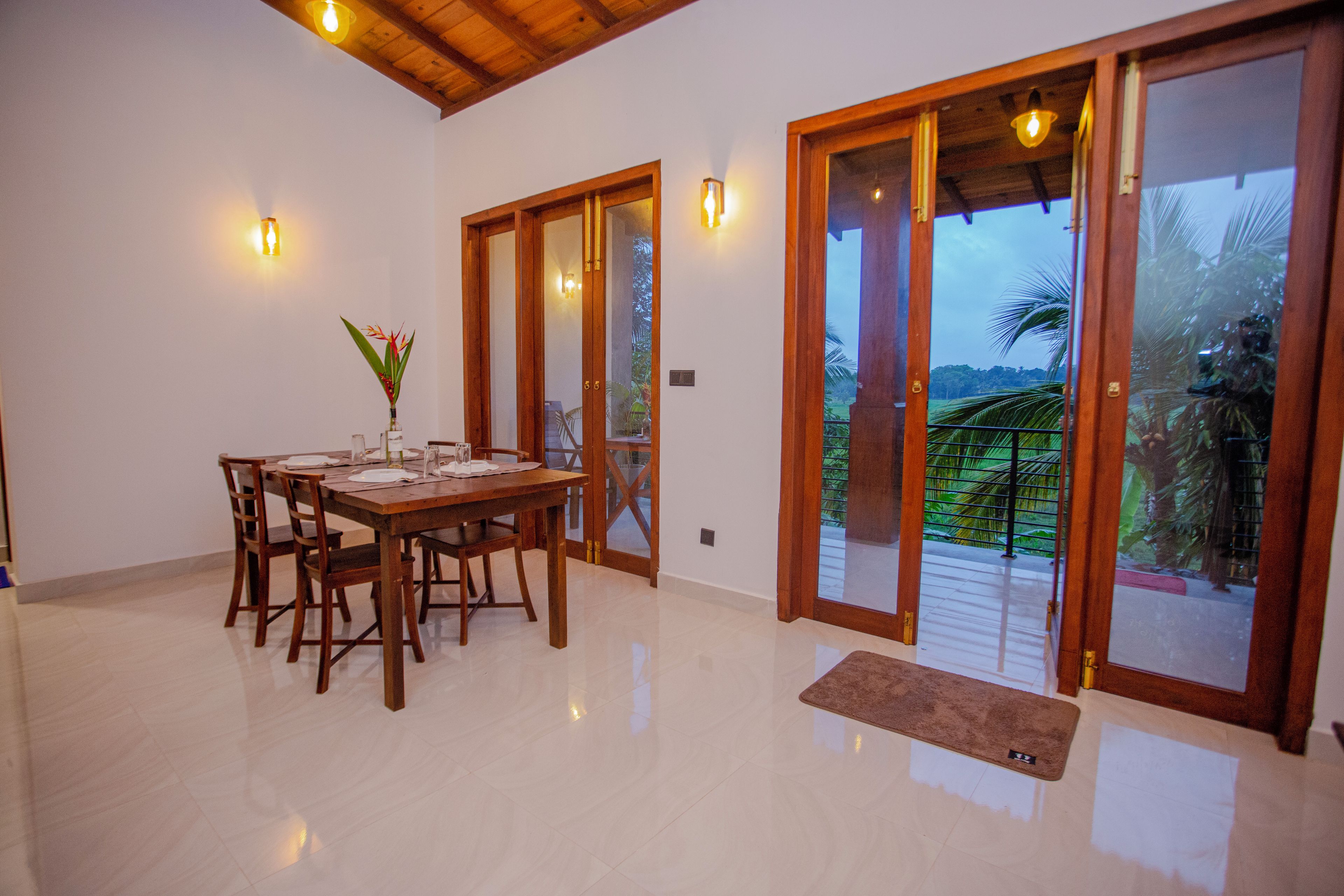 Comedor con mesa de madera dispuesta para cuatro, un florero con flores y vista a un jardín a través de puertas de cristal.