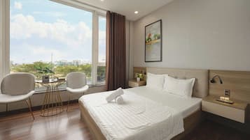 Habitación de hotel con un ventanal grande, una cama con ropa blanca, dos sillas, una mesita de noche con una lámpara y un cuadro en la pared.