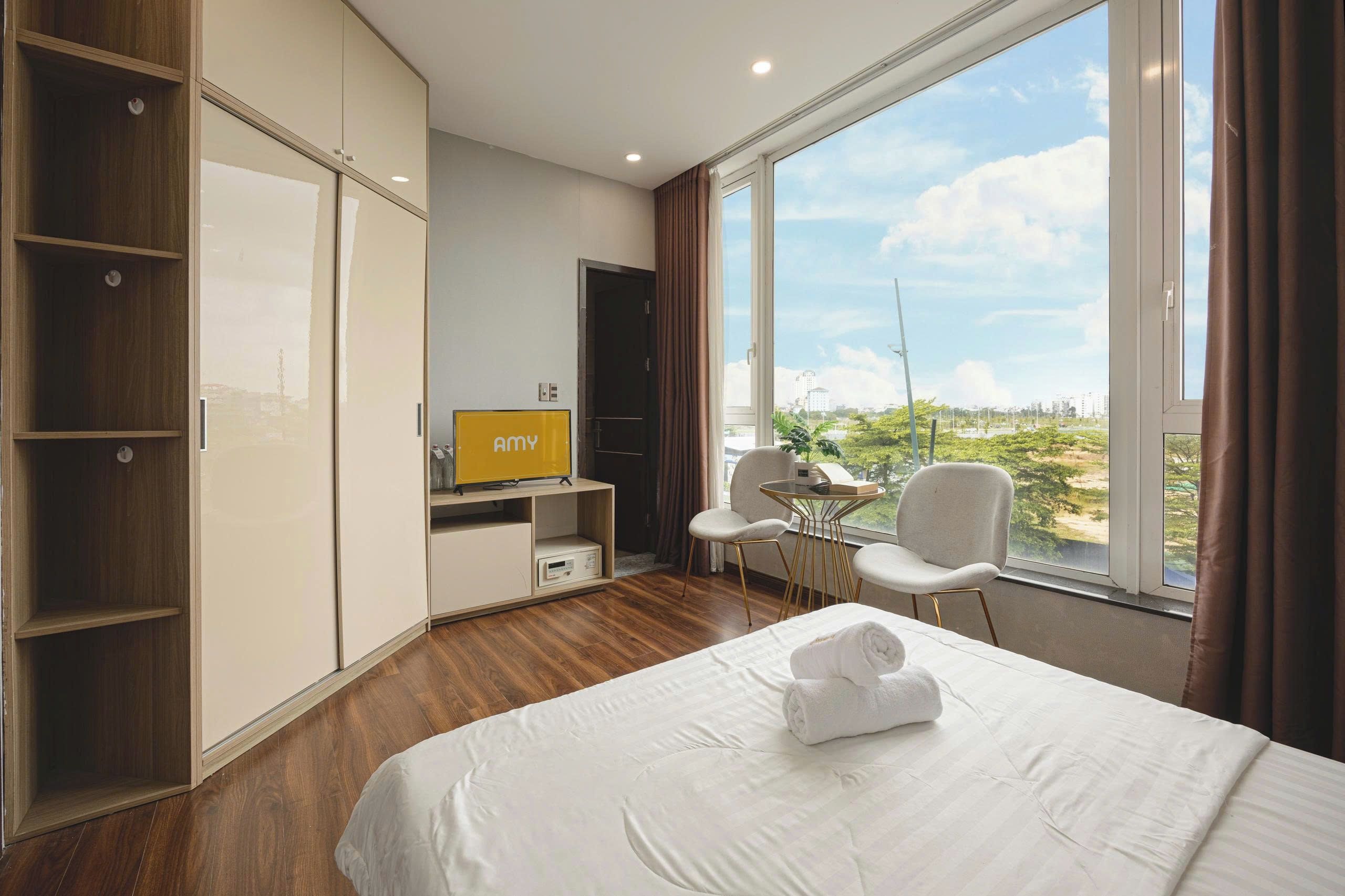 A hotel room with a bed, a TV, a desk with a chair, and a large window offering a view of the city.