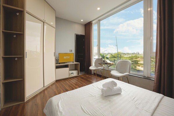 A hotel room with a bed, a TV, a desk with a chair, and a large window offering a view of the city.