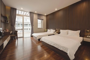 Habitación de hotel con suelo de madera, dos camas, un escritorio, una silla, un televisor y un balcón con cortinas.