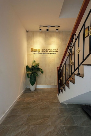 Una escalera con barandilla de madera y balaustradas metálicas, que conduce a la entrada de un apartamento con una planta en maceta y un cartel en la pared que muestra el nombre y los datos de contacto del apartamento.