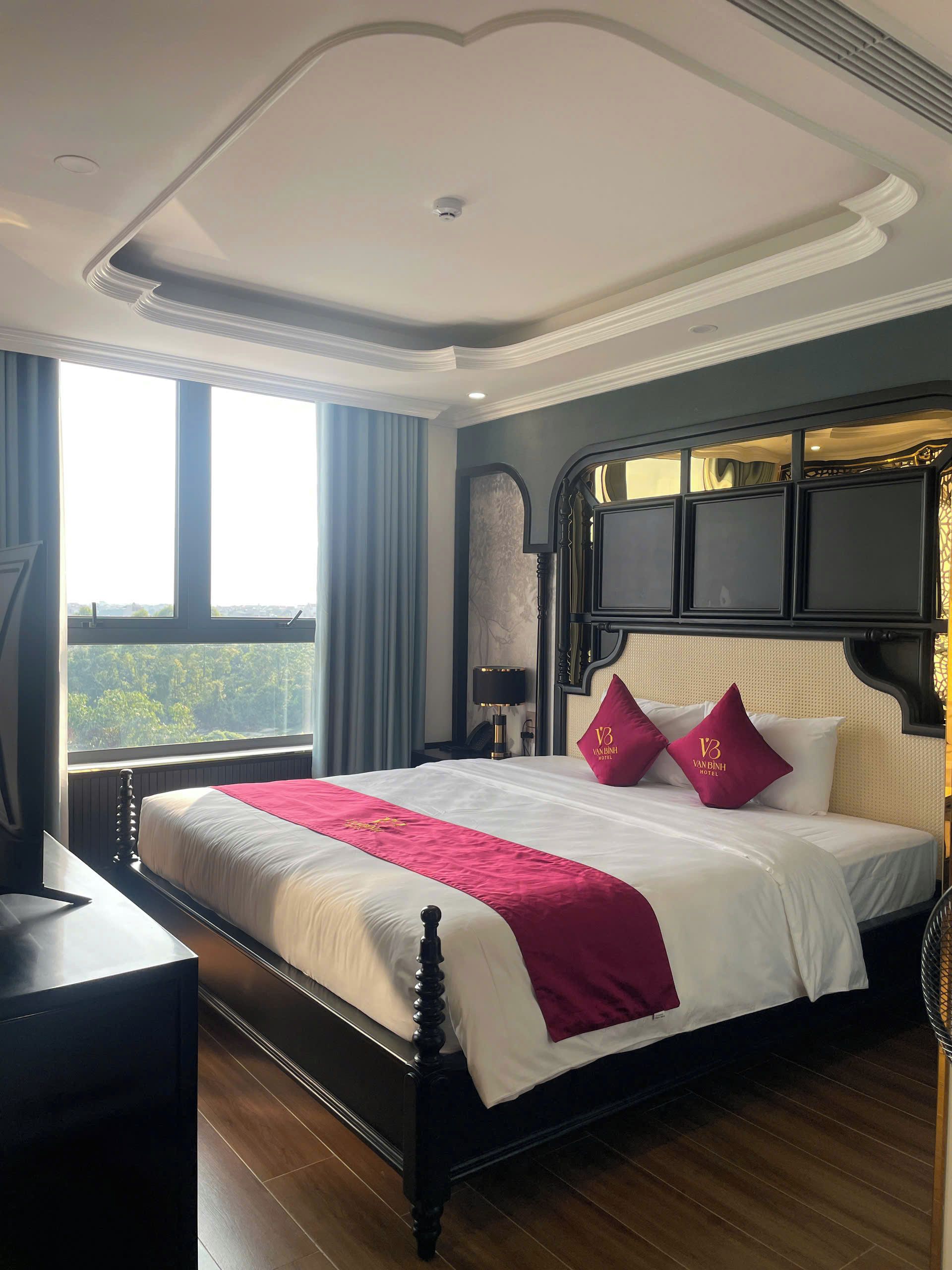 A hotel room with a large bed, a desk, and a view of greenery outside.