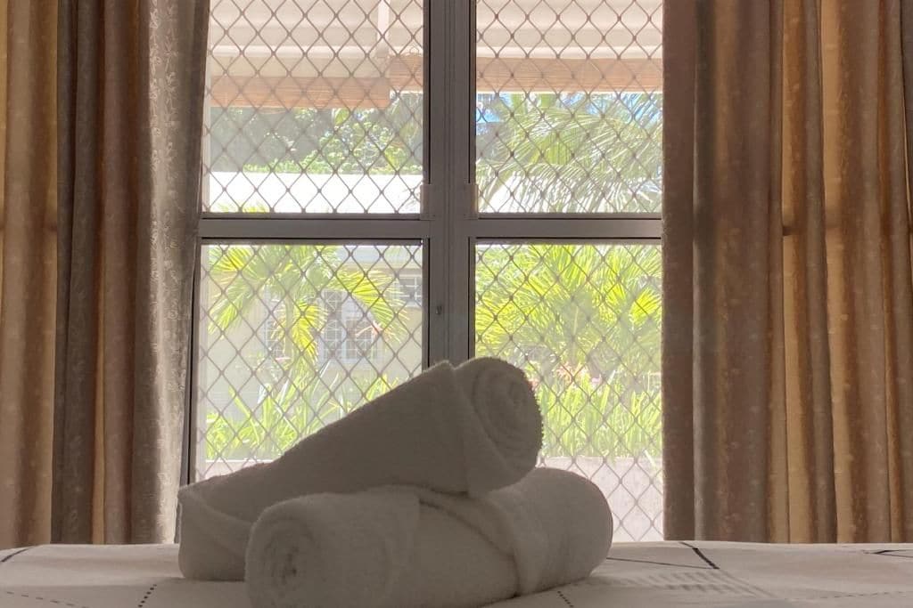 A window with a view of greenery and a building outside, a rolled white towel on a surface, and curtains partially drawn.