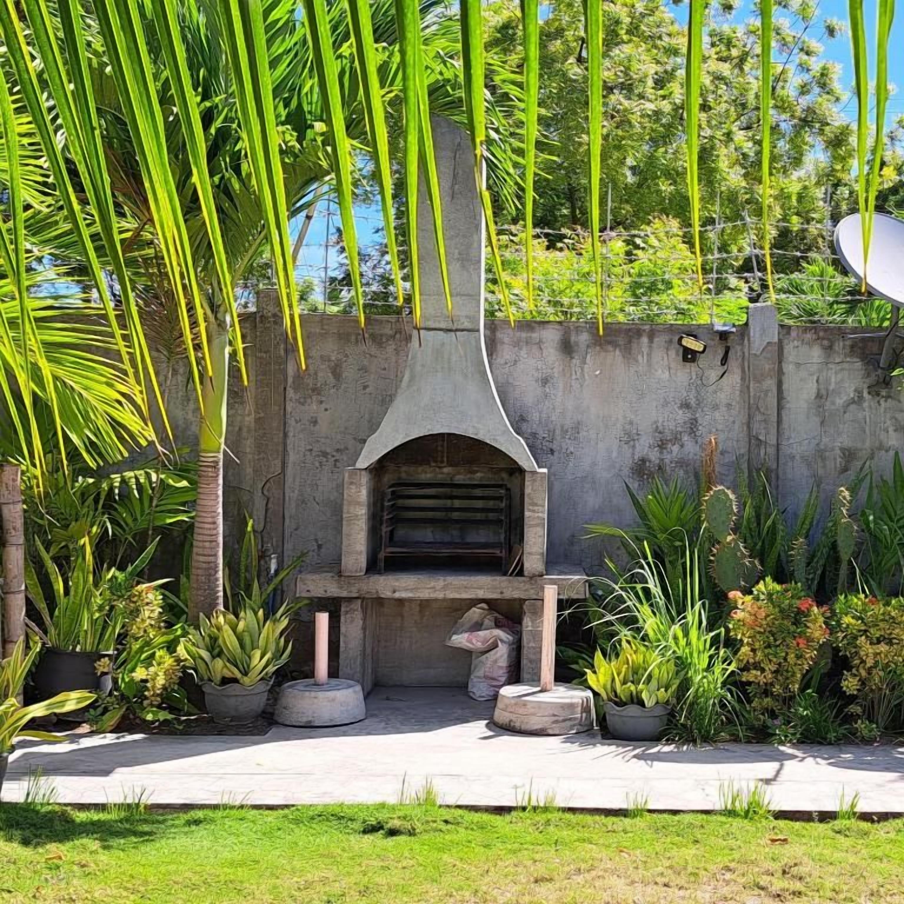 Un espacio exterior de concreto con una parrilla integrada, rodeado de plantas en macetas y follaje variado.
