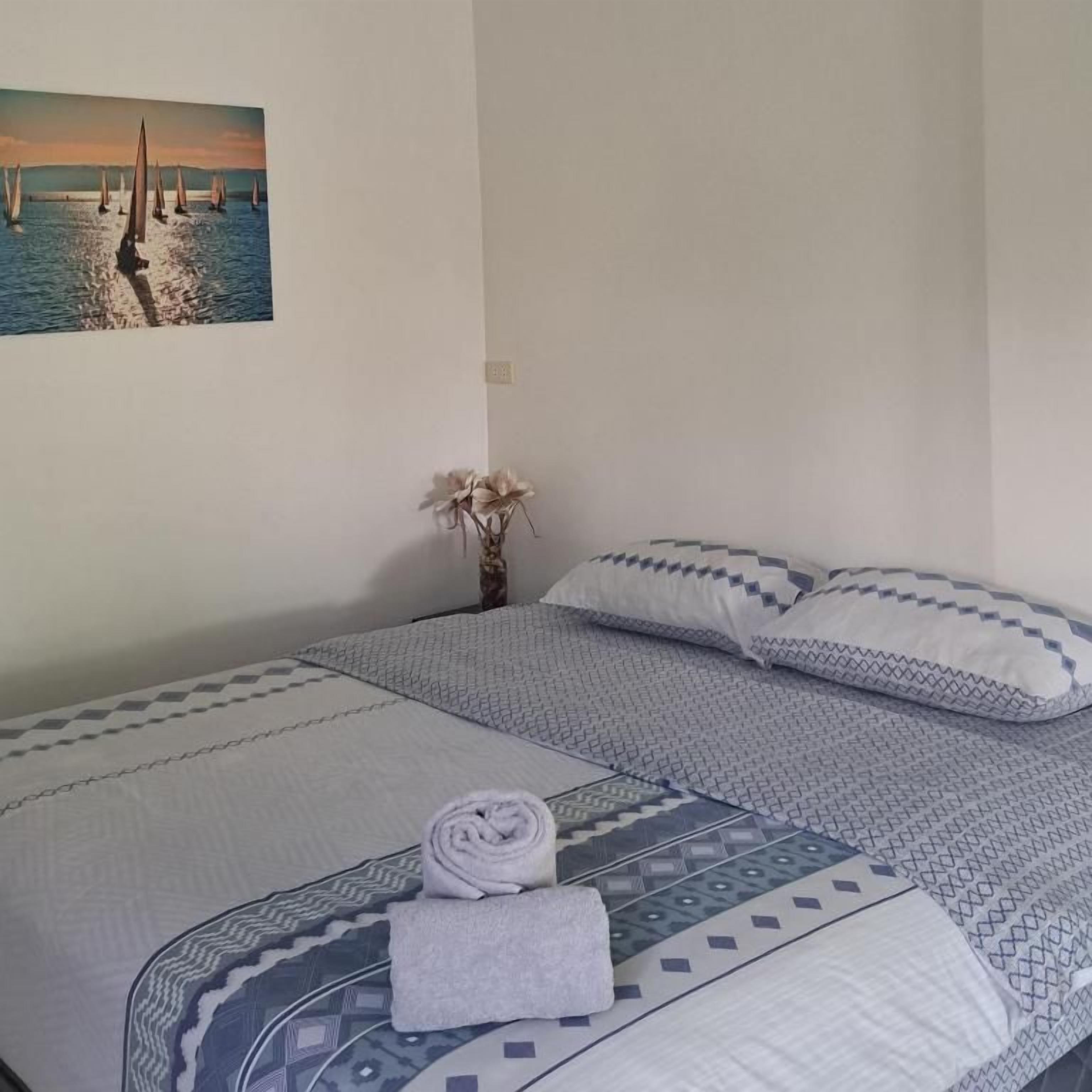 Una cama bien tendida con ropa de cama estampada, una toalla enrollada y un cuadro de veleros en la pared.
