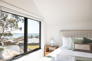 A bedroom with a large window, a bed with pillows, a bedside table with a lamp, and a view of the sea and buildings. - Timbre Blue - Want for nothing (Break O'Day)