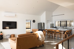 A modern living room with a sofa, TV, and a kitchen area with bar stools. - Timbre Blue - Want for nothing (Break O'Day)