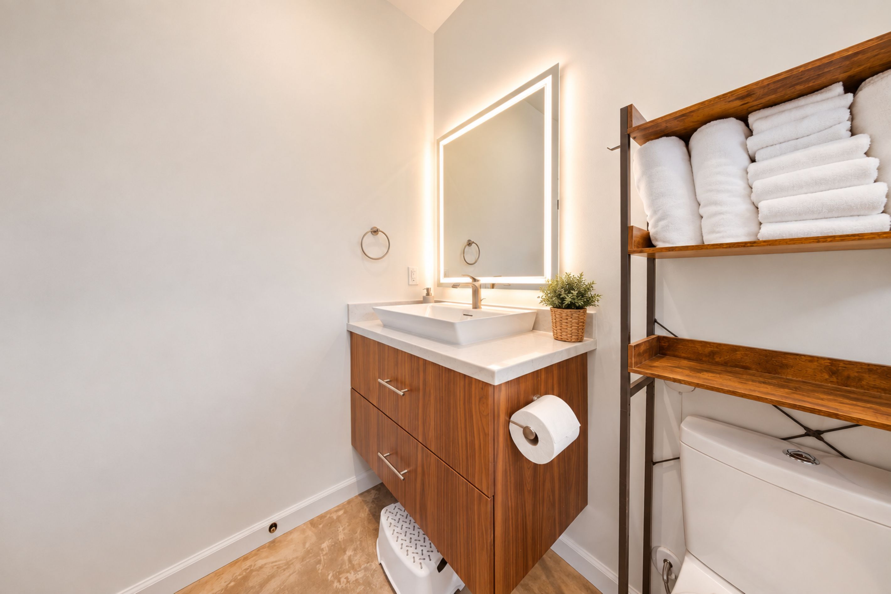 Een badkamer met een houten wastafelmeubel, een witte wasbak, een spiegel met ingebouwde verlichting, een toilet en een plank met opgevouwen handdoeken.