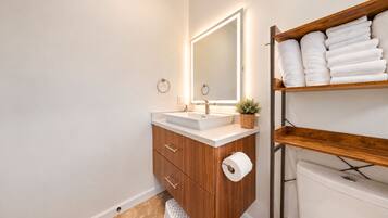 Een badkamer met een houten wastafelmeubel, een witte wasbak, een spiegel met ingebouwde verlichting, een toilet en een plank met opgevouwen handdoeken.