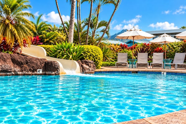 Une piscine avec un toboggan, des chaises longues, des parasols et des plantes tropicales.