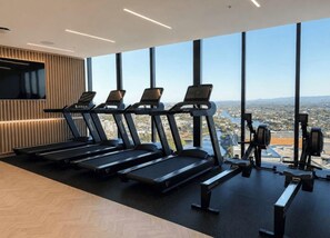 Une salle de sport équipée de tapis de course et d’appareils de musculation, avec de grandes fenêtres offrant une vue sur la ville.