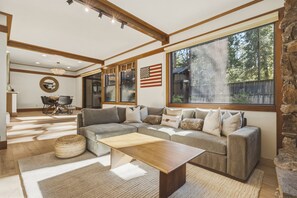 Ein geräumiges Wohnzimmer mit einer Eckcouch, einem Holztisch und einem großen Fenster mit Blick auf Bäume.