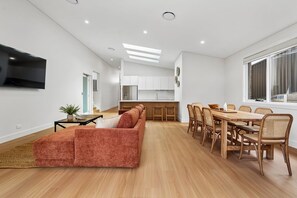 A modern living room with a sofa, dining table, and chairs. There is a kitchen in the background with white cabinets and a flat-screen TV mounted on the wall. - The Boat House - Four Bedroom Coastal Home in Sawtell (Sawtell)