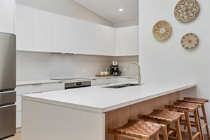 A modern kitchen with white cabinetry, a stainless steel refrigerator, a built-in island with stools, and decorative woven plates on the wall. - The Boat House - Four Bedroom Coastal Home in Sawtell (Sawtell)