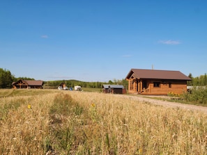 A rustic cabin with a large porch and a covered outdoor area, surrounded by a field and a clear sky.