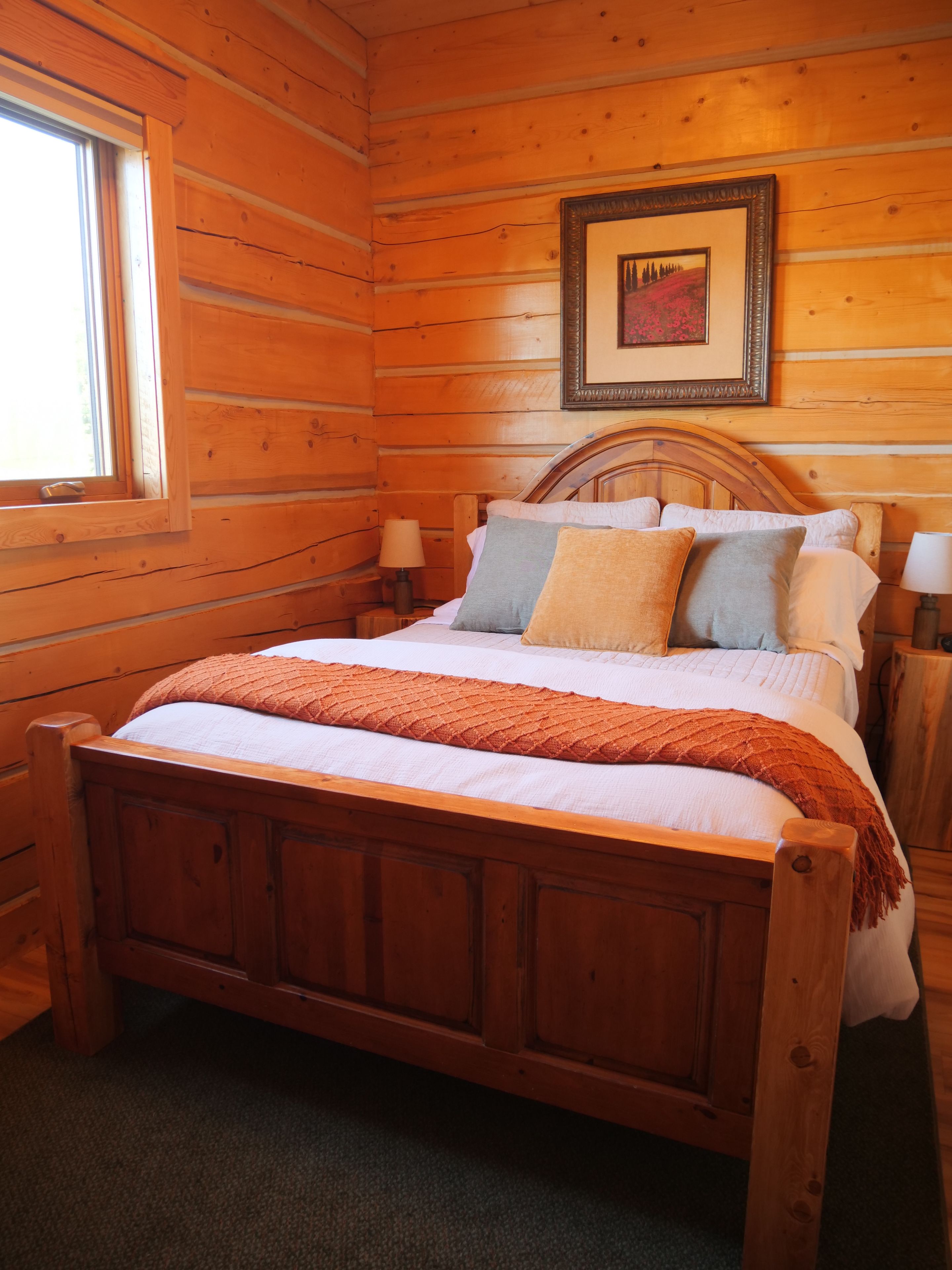 A wooden cabin room with a bed, two bedside tables, a window, and a framed picture on the wall.