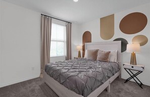 A bedroom with a bed, two bedside tables, a window with curtains, and wall art. - Sunshine Hideaway Near Disney • Resort Pool • Family-Friendly 3BR (Davenport)