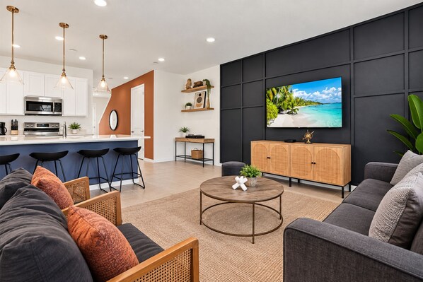 Een moderne woonkamer met een grijze bank, een ronde salontafel en een televisie waarop een tropisch strand te zien is.