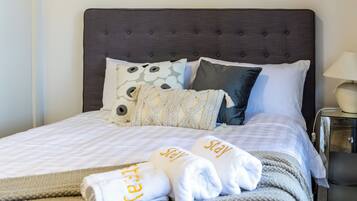 Una cama bien hecha con cabecera gris, ropa de cama blanca y almohada decorativa. Hay un cuadro en la pared sobre la cama.