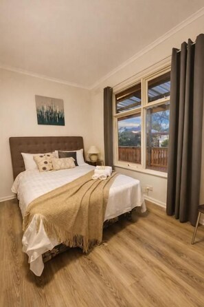 A bedroom with a bed, a nightstand, a window with curtains, and a view of the outdoors.