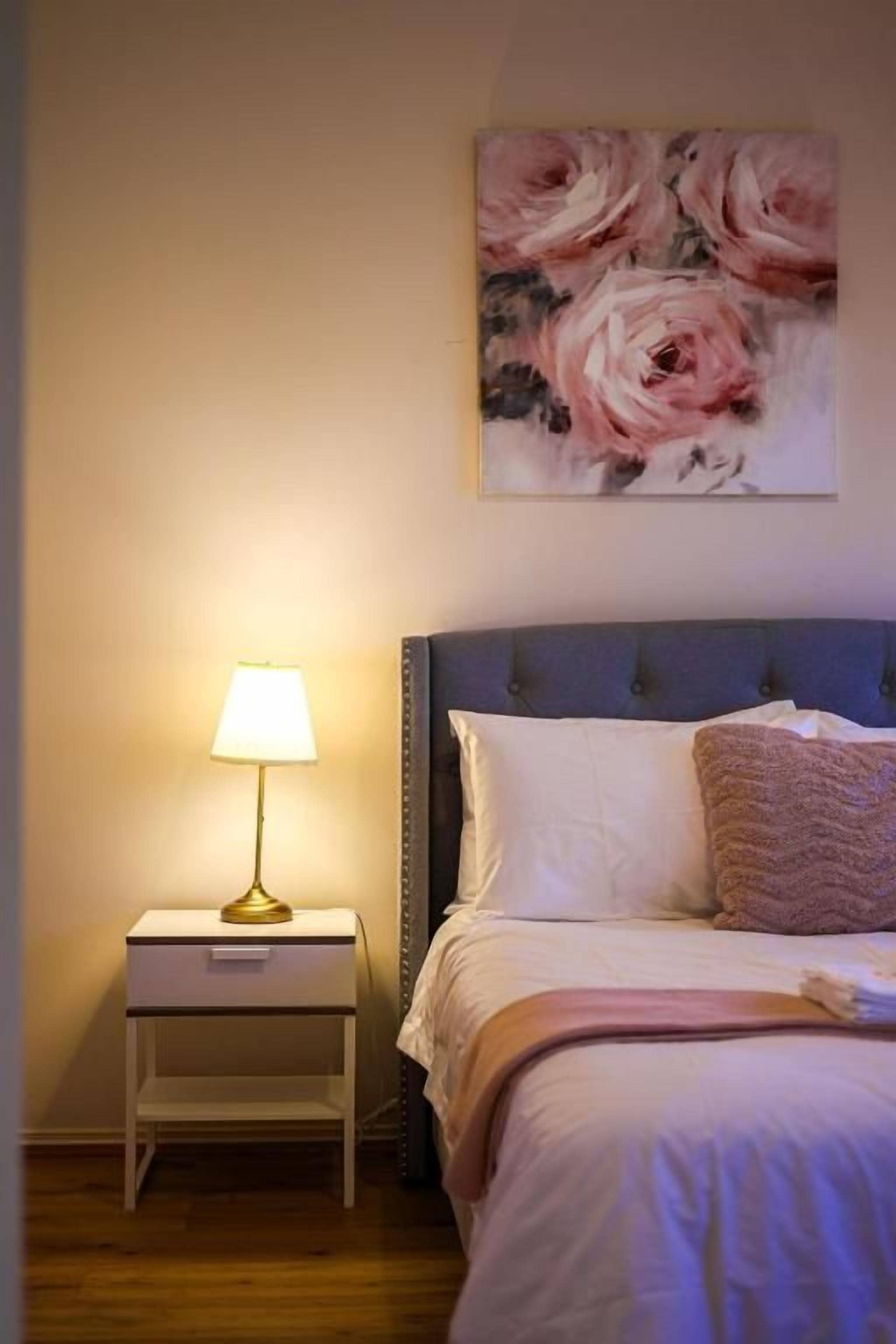 Une chambre à coucher avec un lit, une table de chevet, une lampe et un tableau représentant des fleurs accroché au mur.
