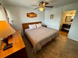 Ein Schlafzimmer mit Bett, Nachttisch, Lampe, Deckenventilator und einem durchgegangenen Tür erkennbaren Badezimmer.