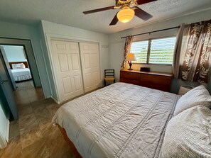 Ein Schlafzimmer mit einem Bett, einer Kommode, einem Fenster mit Vorhängen, einem Deckenventilator und einer Tür zu einem anderen Raum.