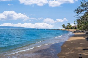 A beach with clear blue water, sandy shore, and a building with a sloped roof. - 2nd Floor Oceanfront 1BD with Panoramic Island Views -Kahana Reef 217 (Lahaina)
