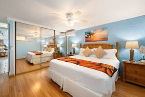 Une chambre d’hôtel avec un grand lit, un ventilateur de plafond, un tableau représentant un coucher de soleil et un miroir sur un des murs.