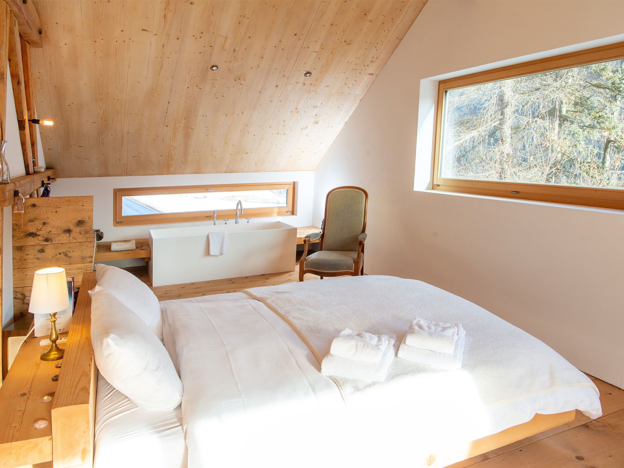 Ein Schlafzimmer mit einem großen Bett, einer Holzbalkendecke, einem Fenster mit Blick auf Bäume, einem Nachttisch mit Lampe und einem Sessel.