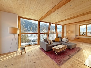 A spacious room with a large window offering a mountain view, a grey sofa, a wooden coffee table, and a red rug.
