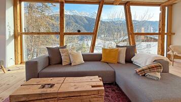 Una sala de estar moderna con un amplio sofá modular, una mesa de centro de madera y vistas a montañas a través de grandes ventanales.