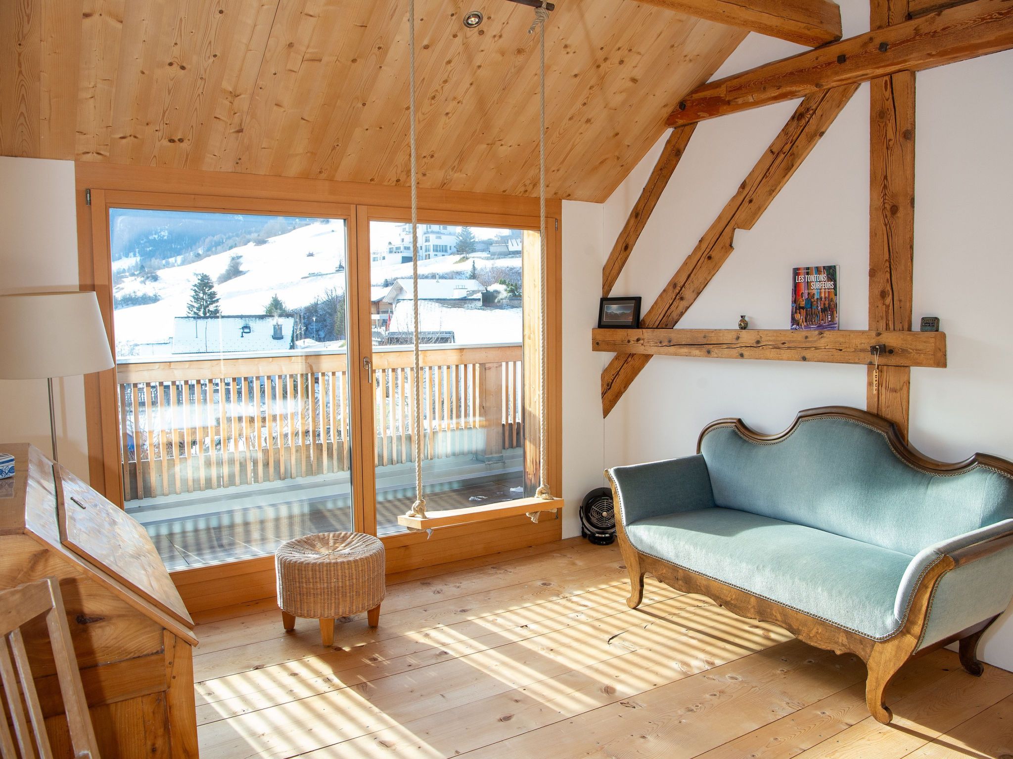 Ein Zimmer mit Holzboden, einer Couch, einem Korbsessel und einem großen Fenster mit Blick auf eine verschneite Landschaft.