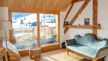 Ein Zimmer mit Holzboden, einer Couch, einem Korbsessel und einem großen Fenster mit Blick auf eine verschneite Landschaft.