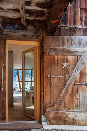 A rustic wooden interior with a large wooden door, a visible outdoor view, and a rope tied to the door.