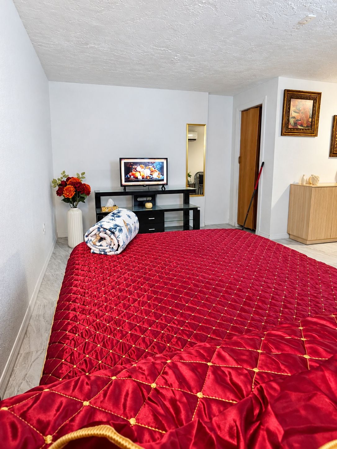 A bedroom with a red quilted bedspread, a television on a black stand, a vase with flowers, and a wooden cabinet.