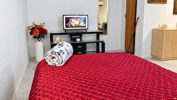 A bedroom with a red quilted bedspread, a television on a black stand, a vase with flowers, and a wooden cabinet.