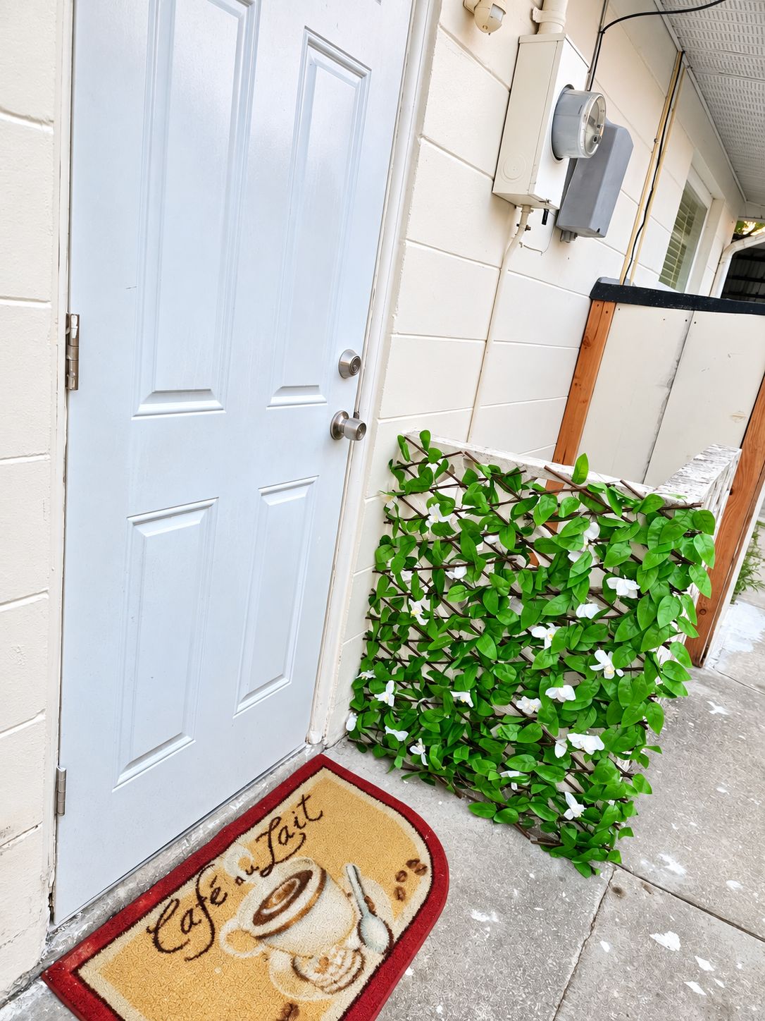 A white door with a doormat that reads 'Café Lait,' a small green plant, and a white wall with a window and an air conditioning unit.