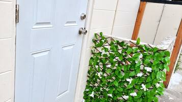 A white door with a doormat that reads 'Café Lait,' a small green plant, and a white wall with a window and an air conditioning unit.