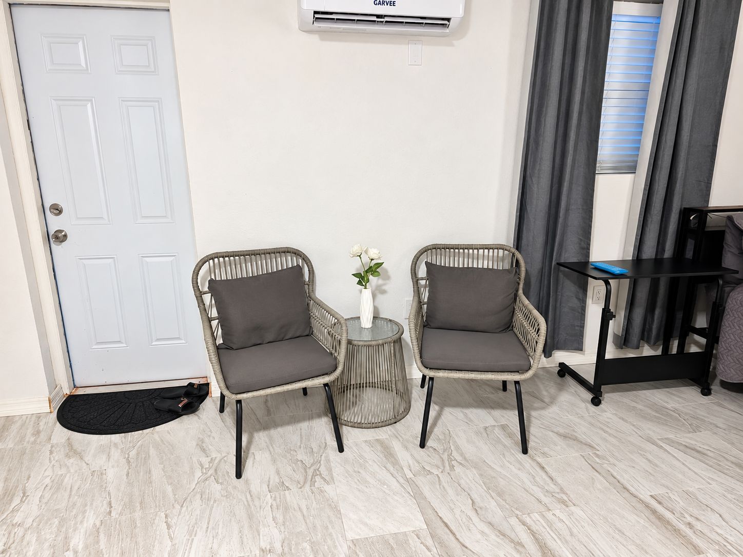 A room with two wicker chairs with cushions, a small round table with a vase of flowers, a white door, and a window with curtains.
