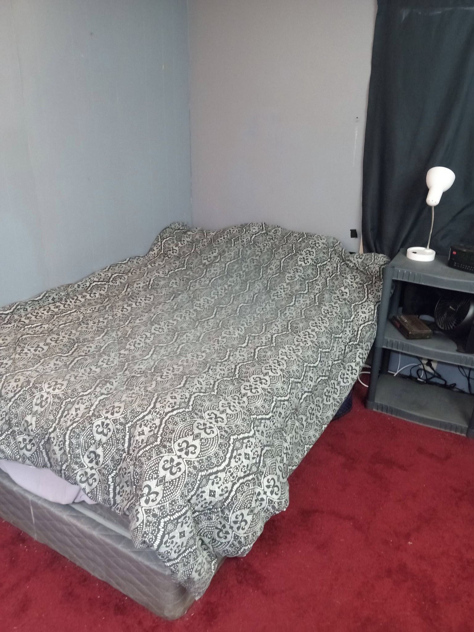 A small room with a single bed, a desk with a lamp, and a shelf with a fan.