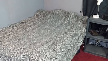 A small room with a single bed, a desk with a lamp, and a shelf with a fan.