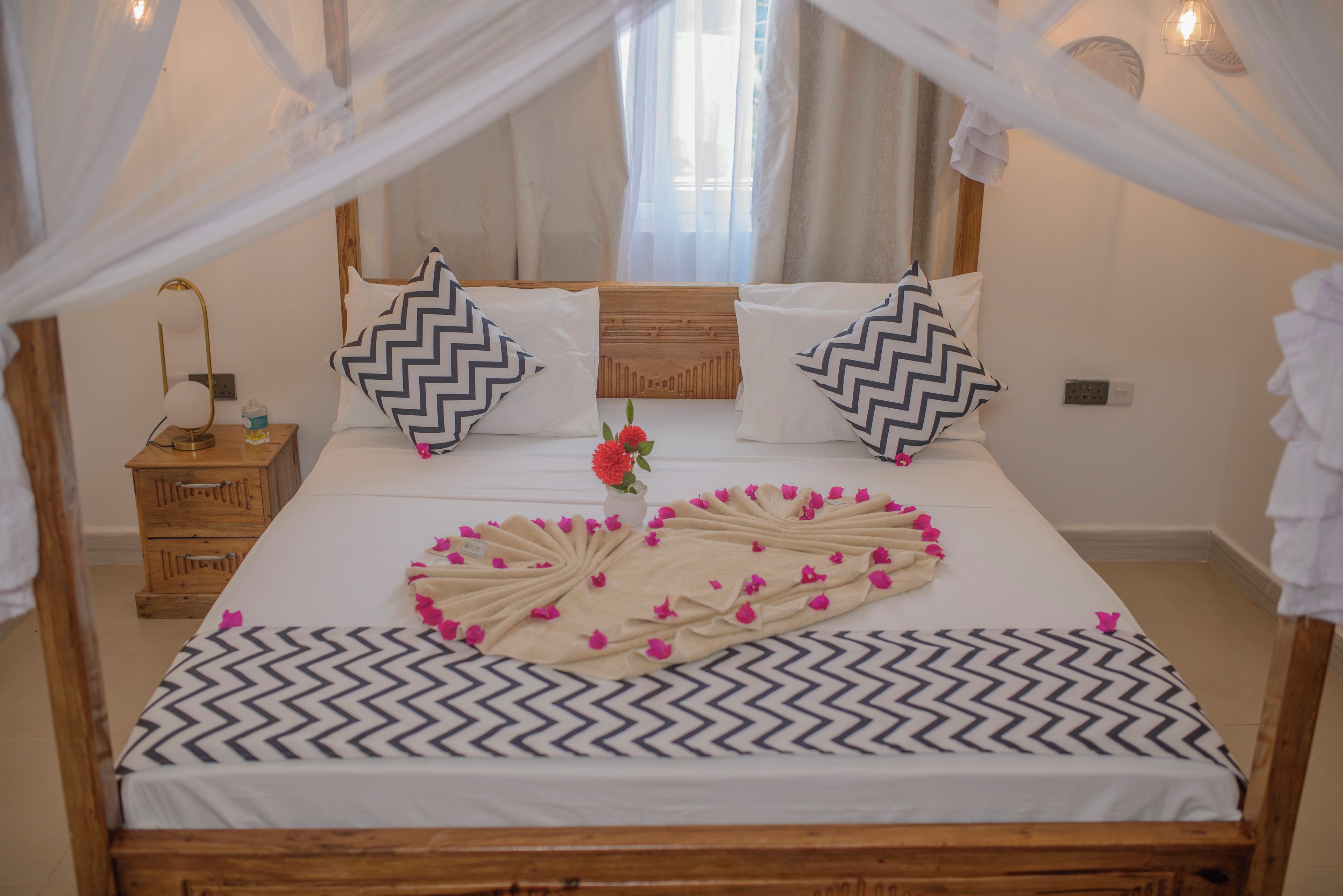 Uma cama com padrão geométrico em branco e preto, duas almofadas com desenho em ziguezague, um criado-mudo de madeira com uma luminária e um vaso com flores.