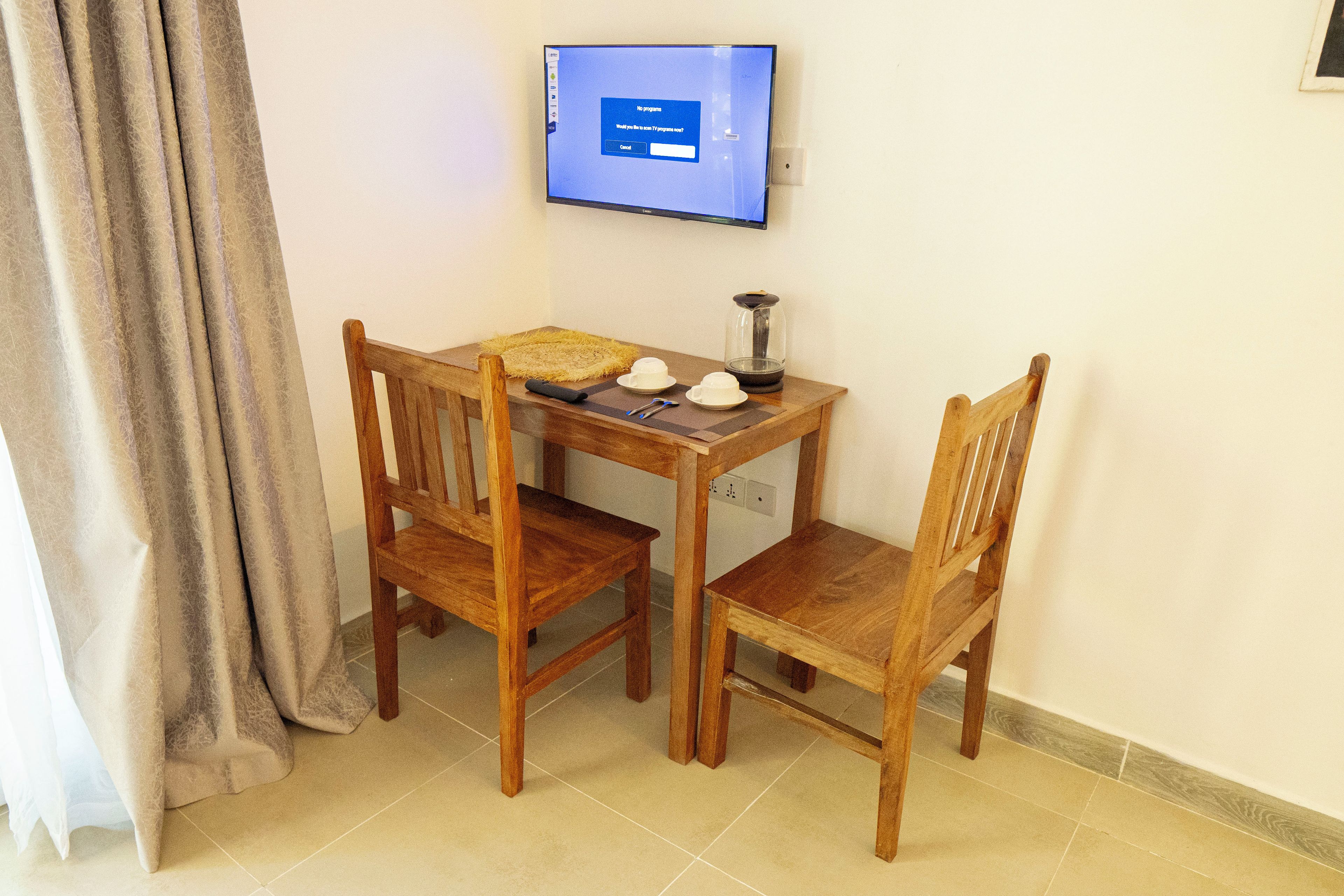 A small wooden table with two chairs, a television mounted on the wall, and a window with curtains.