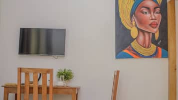 Quarto com cadeira de madeira, mesa pequena, televisão na parede e uma pintura.