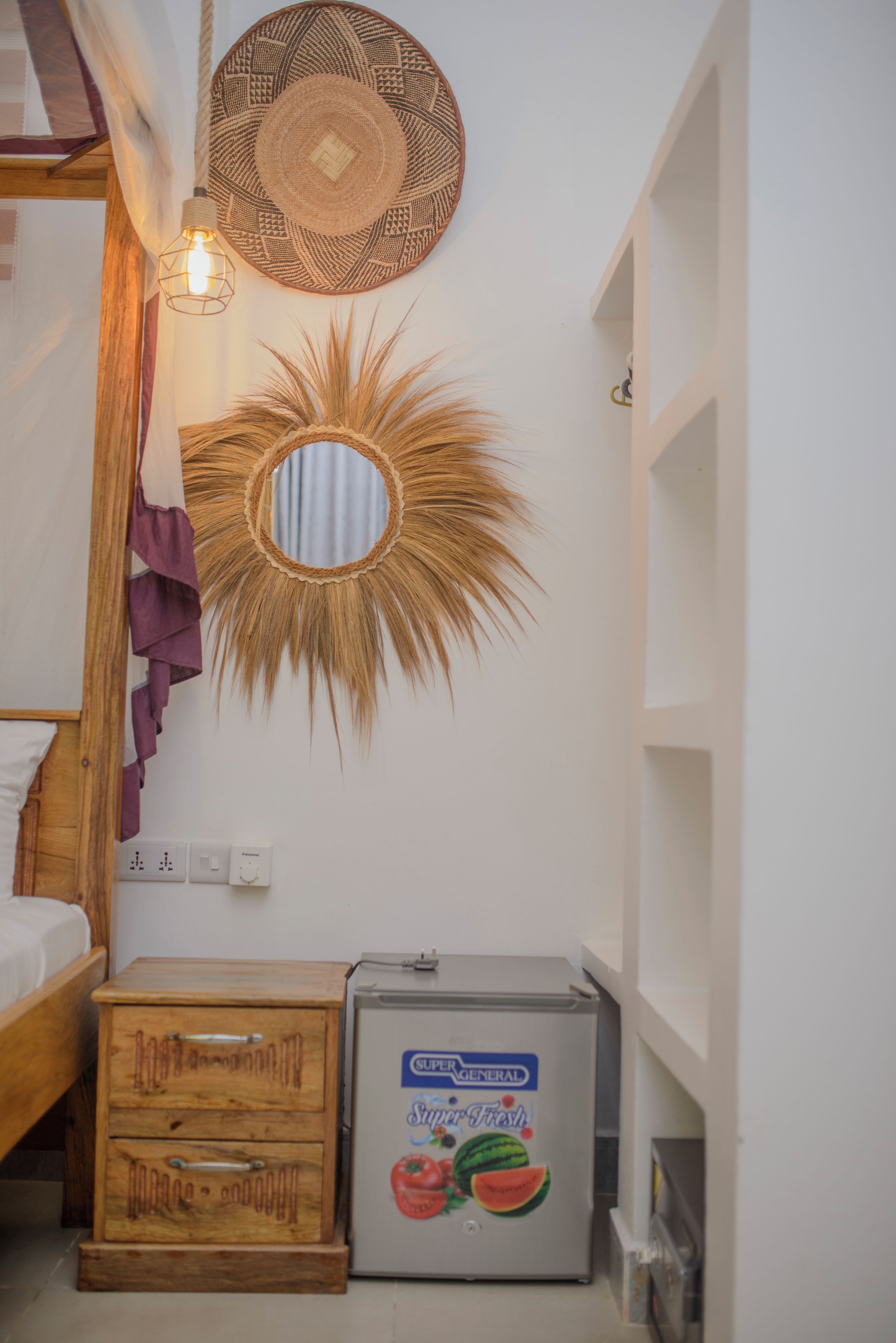 Un dormitorio con una cama, un aparador de madera, un refrigerador, un espejo montado en la pared y un espejo decorativo con forma de sol.