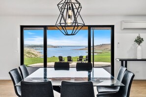 Ein Essbereich mit einem Glastisch und Stühlen, einer geometrischen Pendelleuchte sowie Blick auf Strand und Meer durch große Glasüren.