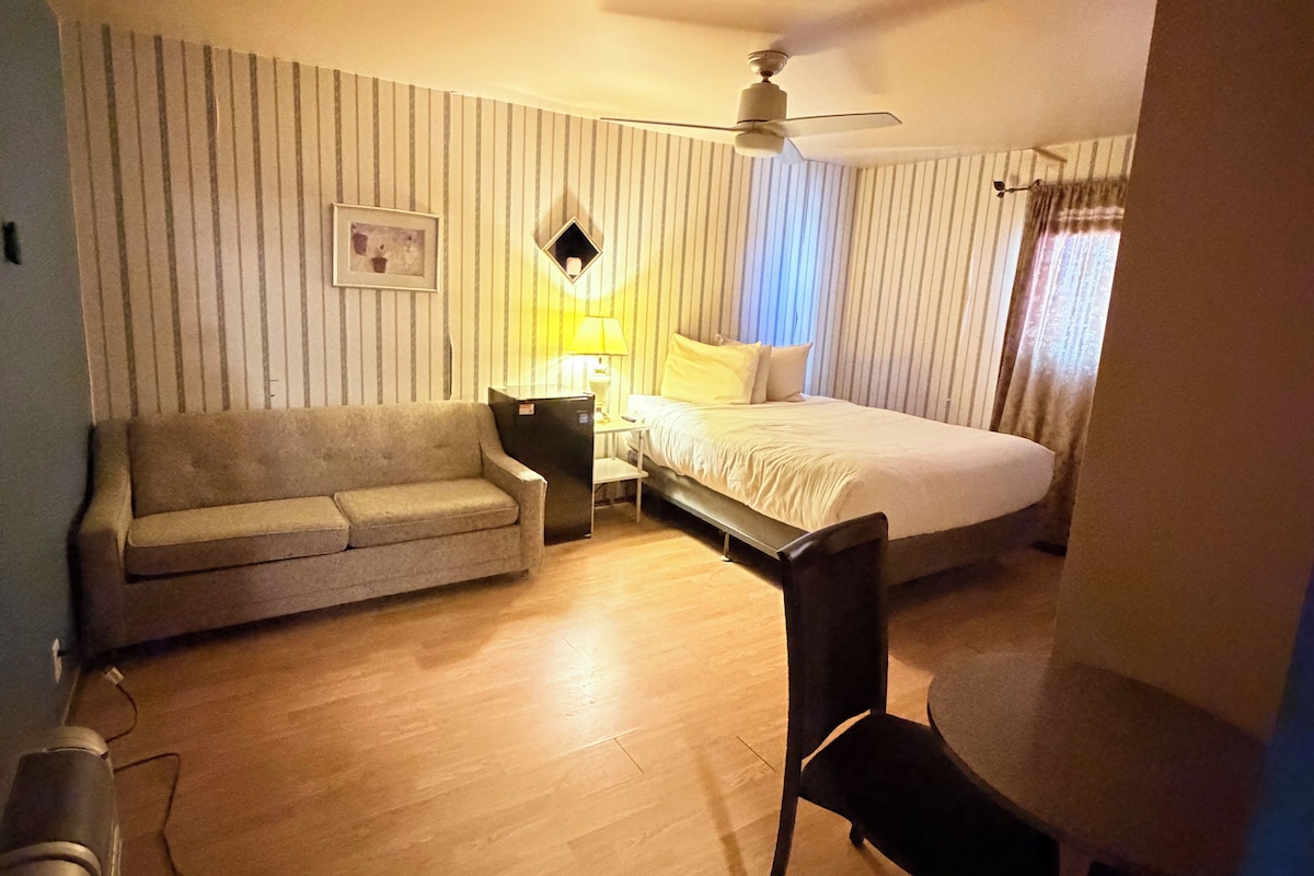 Hotelkamer met een bed, bureau, stoel, bank, nachtkastje, lamp, plafondventilator en raam met gordijnen.